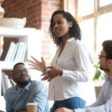 Ambitious smart african black female employee speaking at diverse meeting share creative idea opinion at group briefing while jealous envious skeptical male coworkers looking listening to colleague