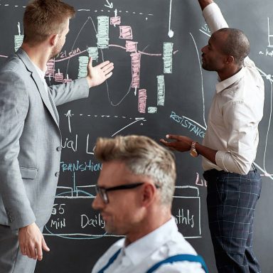 Middle-aged trader looking at computer screen while working in office. Diverse employees standing near blackboard full of charts in background. Stock trading, people, business concept. Vertical shot