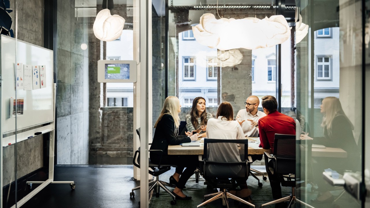 En gruppe mennesker sidder ved et bord under et forretningsmøde i et lyst, moderne kontor. Teamet taler forretning, mens pie diagrammer kan ses hængende på væggen. Store vinduer ses i baggrunden.