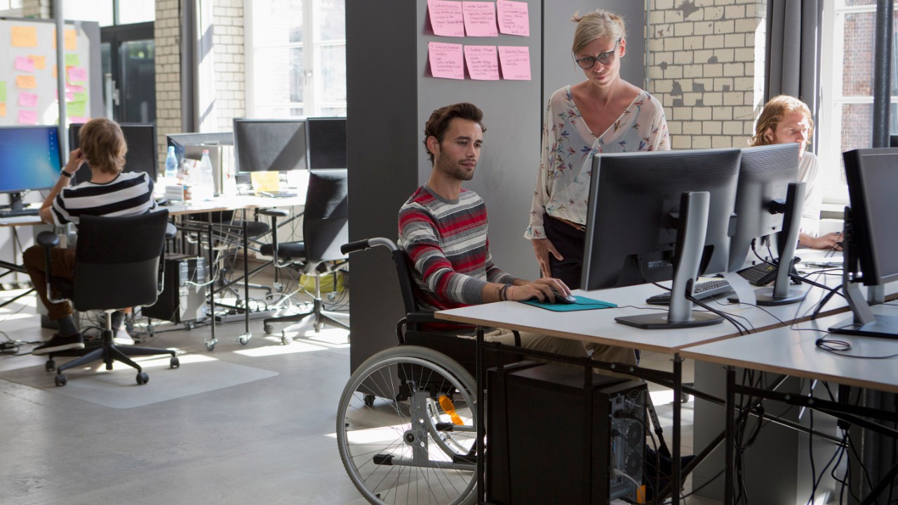 Kontorscene med personer, der arbejder på computere, herunder én person i en kørestol