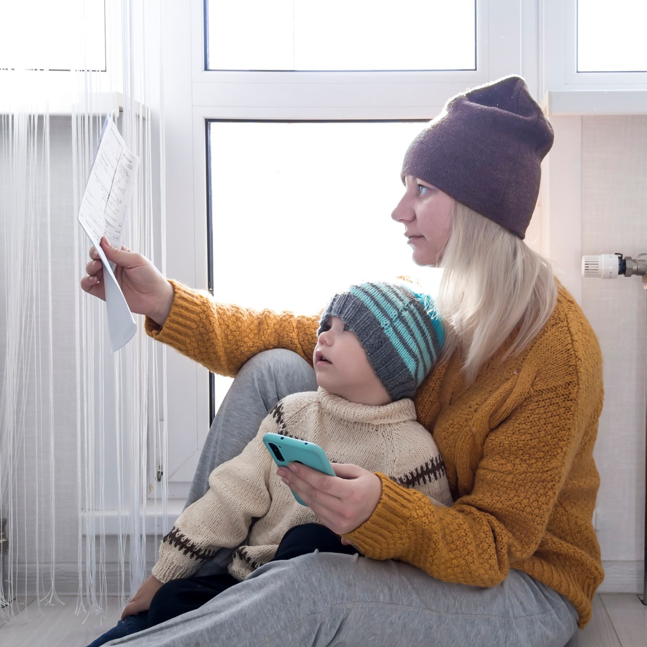 .Una madre con un niño pequeño con un suéter amarillo y sombreros está contando dinero y pensando en cómo pagar facturas e impuestos, cerca de un calentador con un termostato.