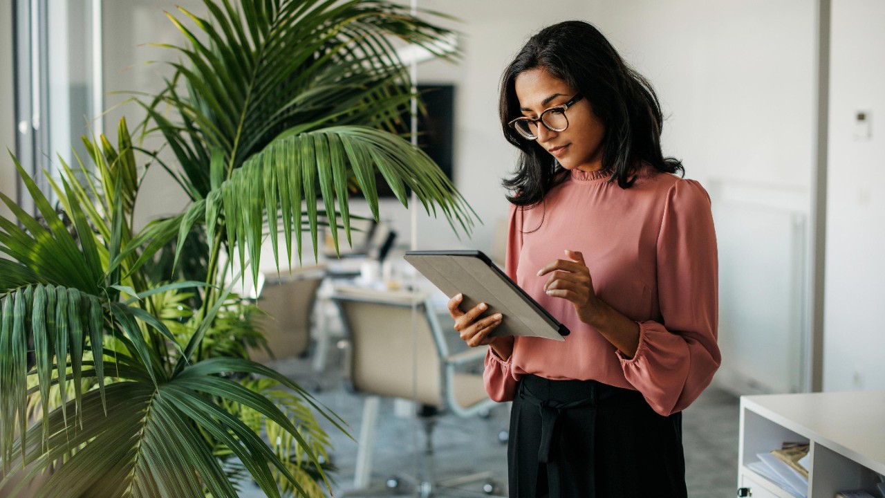 Vrouwelijke leidinggevende met halflang zwart haar draagt een bril en zakelijke kleding die buiten de moderne vergaderruimte staat en gegevens op een tablet controleert.